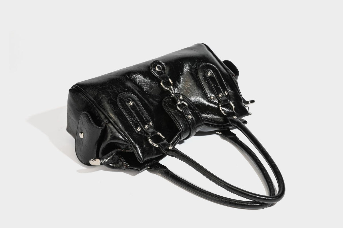 Glänzende, geknautschte Barrel-Satchel mit zwei oberen Tragegriffen, vorderem Klappen-Riemen-Panel, Ring- und Nietenbeschlägen sowie einem abnehmbaren