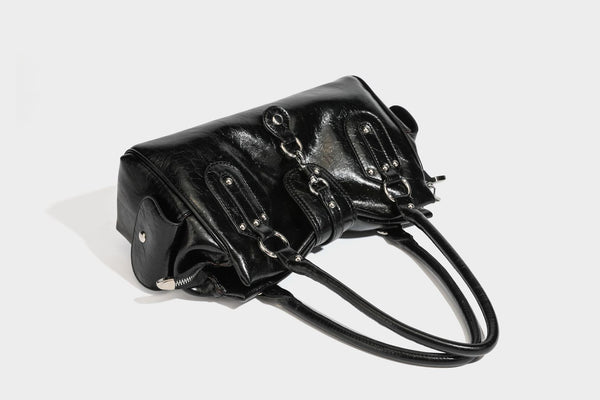 Glänzende, geknautschte Barrel-Satchel mit zwei oberen Tragegriffen, vorderem Klappen-Riemen-Panel, Ring- und Nietenbeschlägen sowie einem abnehmbaren
