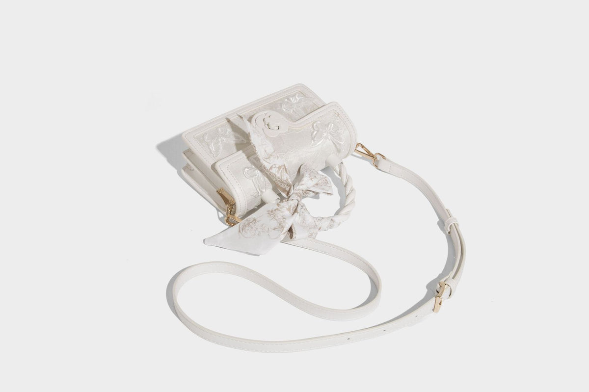 Small structured white flap bag with butterfly embroidery, flower-shaped turn-lock clasp, braided top handle with scarf bow