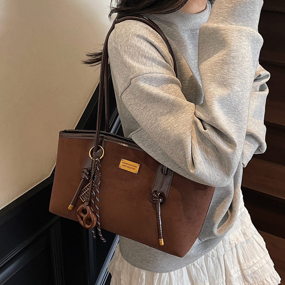 Structured suede-finish tote bag with glossy black leather-look trim, dual top handles, and decorative hanging charm in