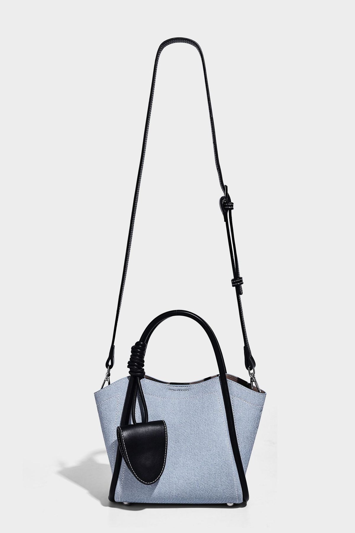 Light blue denim trapezoid tote bag with black leather-look trim, knotted top handles, detachable crossbody strap, front