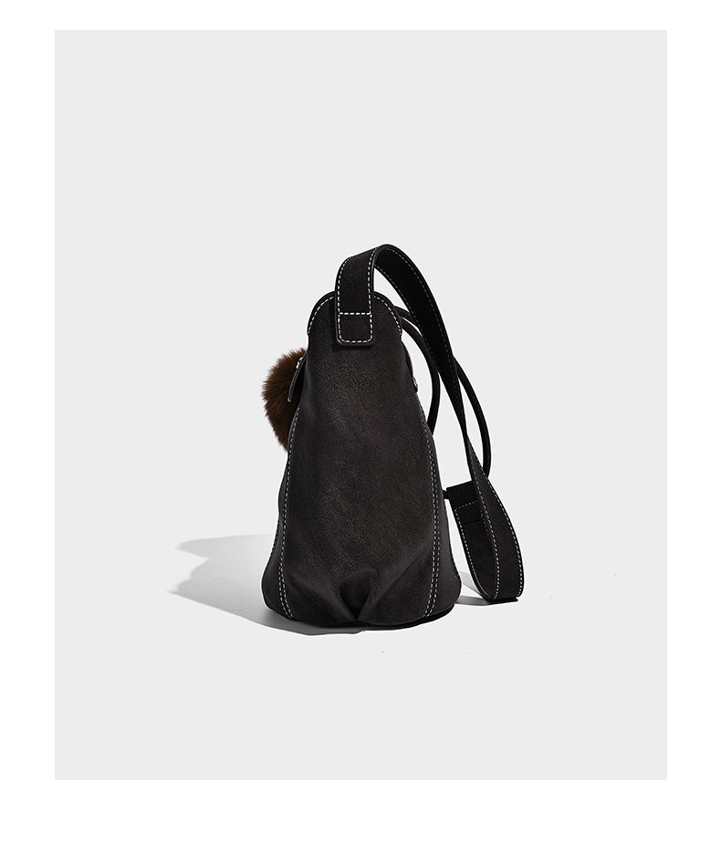 Small matte textured bucket crossbody bag with flap closure, gold-tone bar hardware, contrast stitching, and pompom tassel