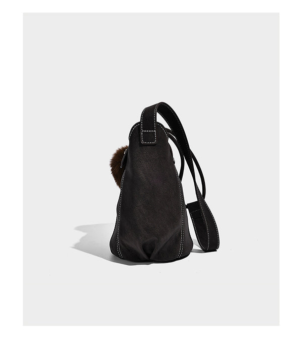 Small matte textured bucket crossbody bag with flap closure, gold-tone bar hardware, contrast stitching, and pompom tassel