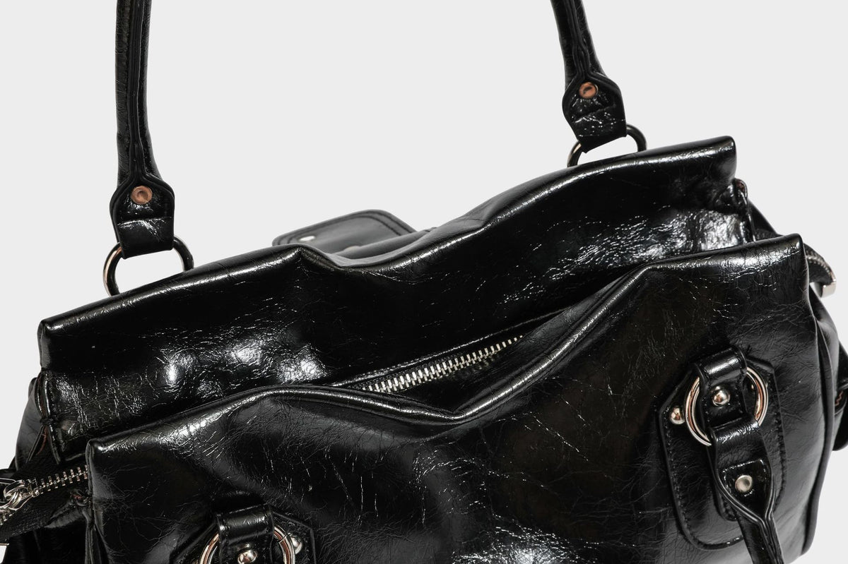 Glänzende, geknautschte Barrel-Satchel mit zwei oberen Tragegriffen, vorderem Klappen-Riemen-Panel, Ring- und Nietenbeschlägen sowie einem abnehmbaren