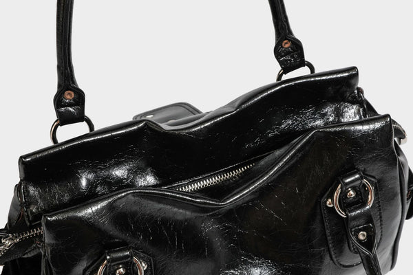 Glänzende, geknautschte Barrel-Satchel mit zwei oberen Tragegriffen, vorderem Klappen-Riemen-Panel, Ring- und Nietenbeschlägen sowie einem abnehmbaren