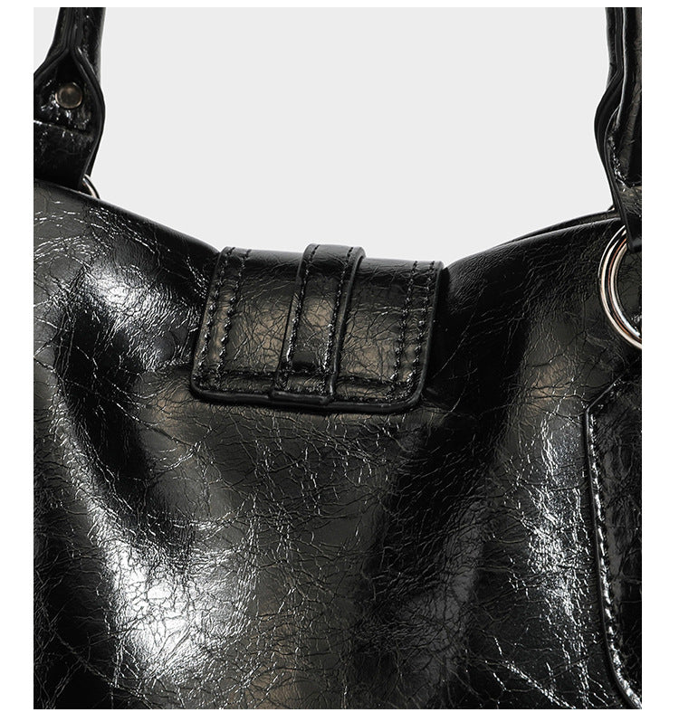 Glänzende, geknautschte Barrel-Satchel mit zwei oberen Tragegriffen, vorderem Klappen-Riemen-Panel, Ring- und Nietenbeschlägen sowie einem abnehmbaren