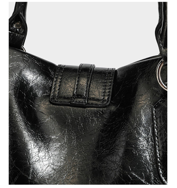 Glänzende, geknautschte Barrel-Satchel mit zwei oberen Tragegriffen, vorderem Klappen-Riemen-Panel, Ring- und Nietenbeschlägen sowie einem abnehmbaren