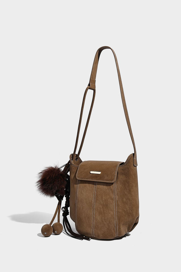 Small matte textured bucket crossbody bag with flap closure, gold-tone bar hardware, contrast stitching, and pompom tassel