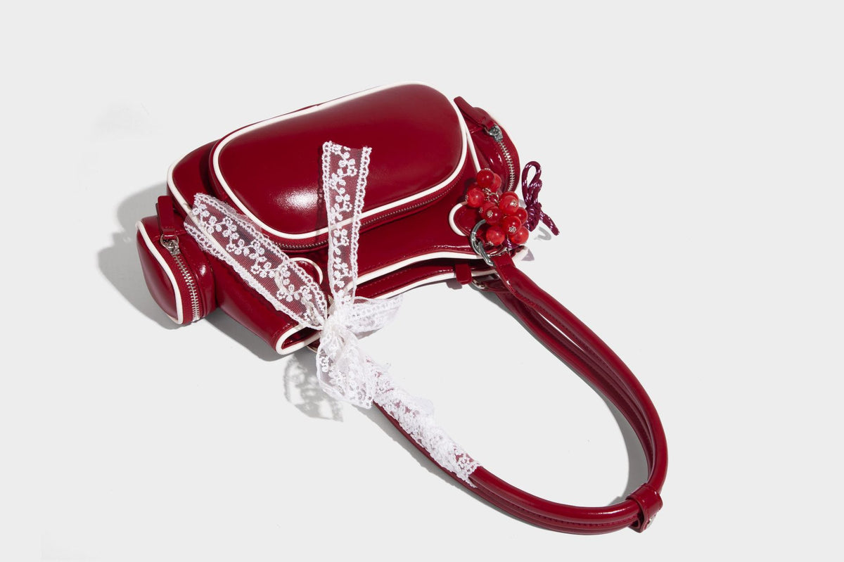 Kleine, formstabile, glänzende Schultertasche mit weißer Paspelierung, Spitzenschleife und Anhänger mit roter Perle, seitlichen Reißverschlusstaschen und silberfarbenem