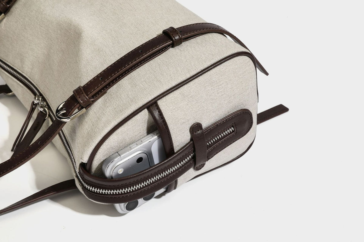 Beige canvas barrel bag with dark brown leather-look trim, silver-tone hardware, top zip closure, and adjustable shoulder