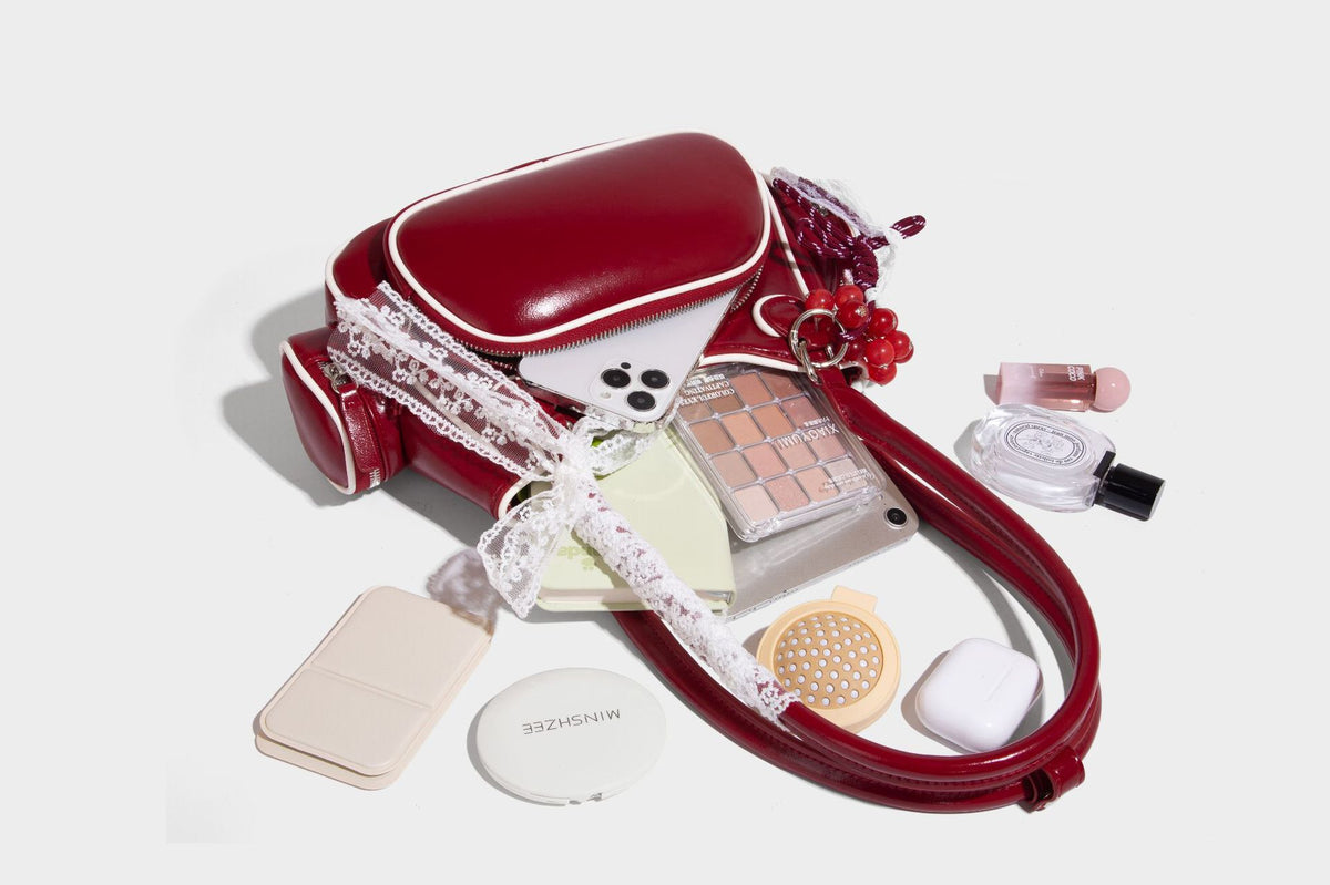 Kleine, formstabile, glänzende Schultertasche mit weißer Paspelierung, Spitzenschleife und Anhänger mit roter Perle, seitlichen Reißverschlusstaschen und silberfarbenem