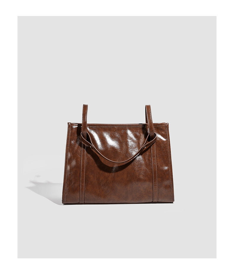 Large structured glossy leather-look tote bag with dual top handles and long shoulder strap in burnished brown, detail view