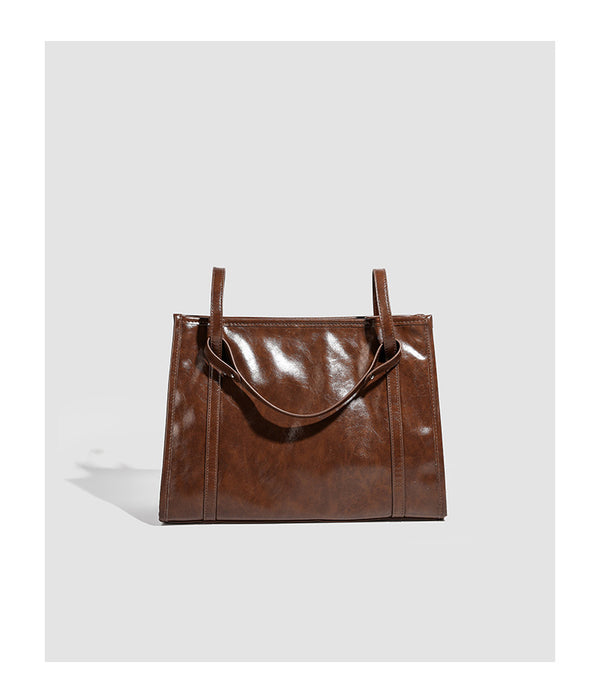 Large structured glossy leather-look tote bag with dual top handles and long shoulder strap in burnished brown, detail view
