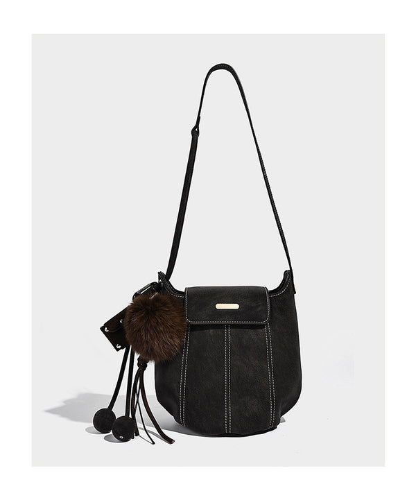 Small matte textured bucket crossbody bag with flap closure, gold-tone bar hardware, contrast stitching, and pompom tassel