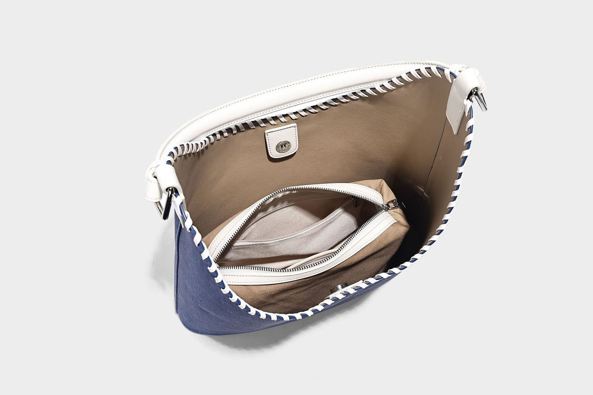 Denim-blue hobo tote with white leather-look shoulder strap, contrast whipstitch trim along curved open top, and silver-tone