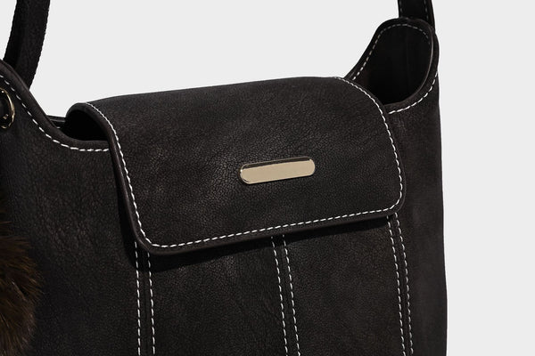 Small matte textured bucket crossbody bag with flap closure, gold-tone bar hardware, contrast stitching, and pompom tassel