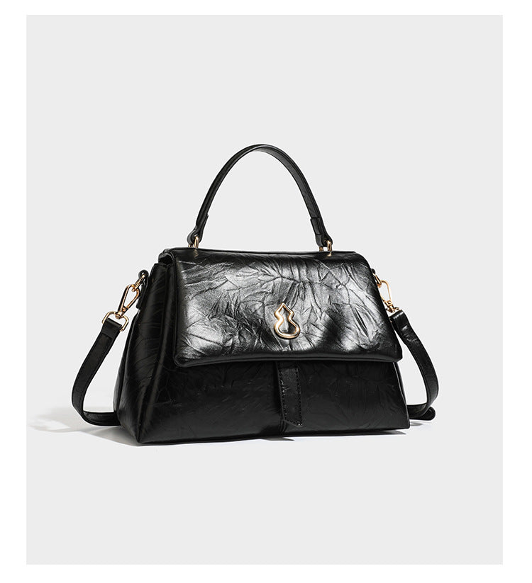 Structured black satchel bag with glossy crinkled leather-look finish, gold-tone emblem flap closure, slim top handle, and