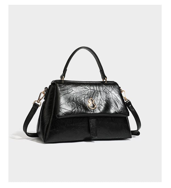 Structured black satchel bag with glossy crinkled leather-look finish, gold-tone emblem flap closure, slim top handle, and