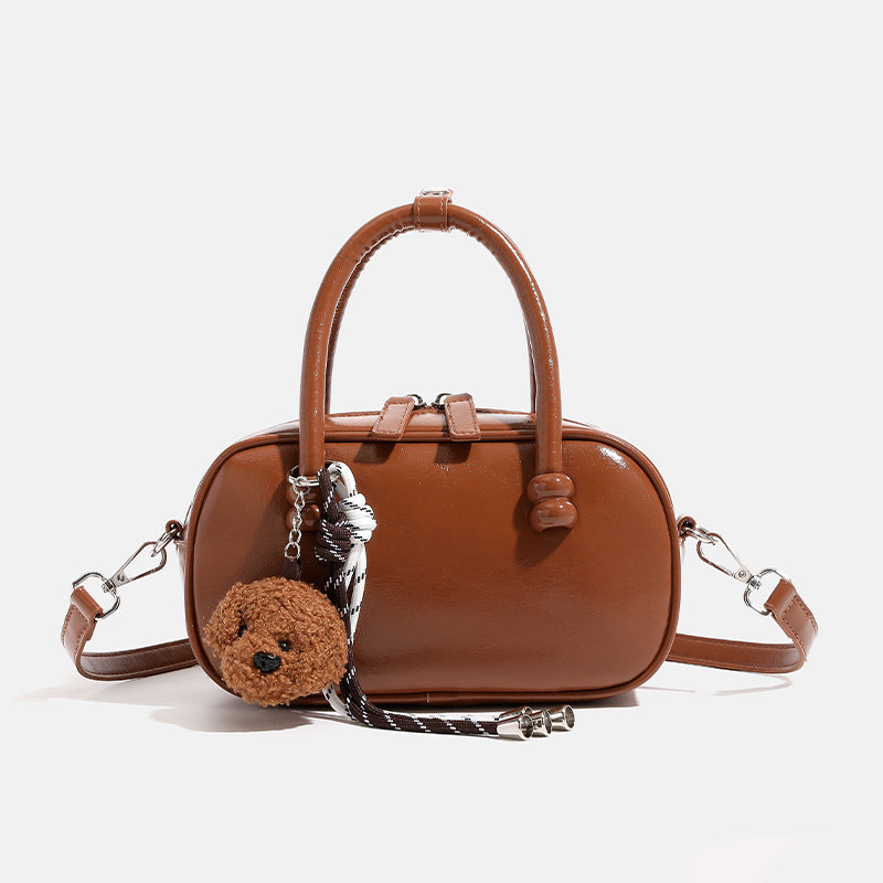 Glossy leather-look mini bowling barrel bag with dual top handles, detachable braided strap, and teddy bear charm, front view