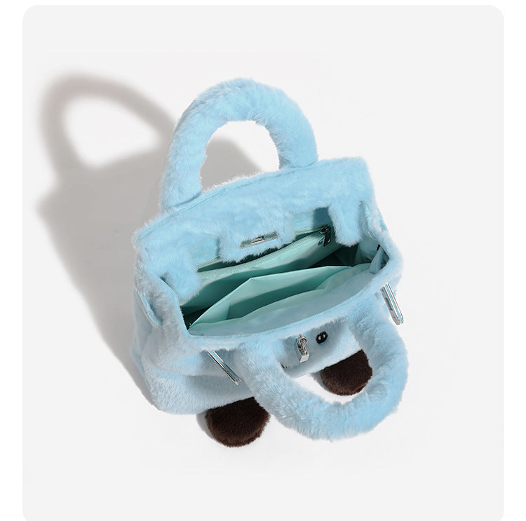 Ice-blue plush faux-fur Kelly-style convertible bag with embroidered smiling face, dark pom-pom feet, silver-tone clasp