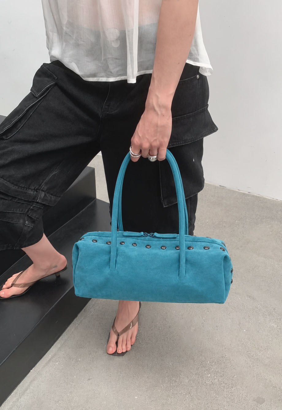 Rechteckige, zylinderförmige Schultertasche mit kuppelförmigen Nieten in Gunmetal-Optik entlang der oberen Naht, zwei flachen Riemen mit Knotendetail und matter Oberfläche, Detail