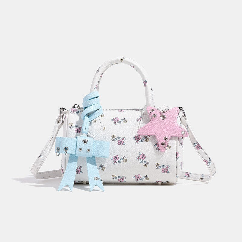 Weiße Mini-Boston-Handtasche mit genarbter Oberfläche und Blumen- oder Kirschprint, pastellblauem Schleifenanhänger, rosafarbenem Sternanhänger und zwei oberen Henkeln