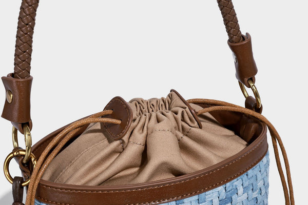 Small light blue woven bucket bag with brown leather-look trim, braided top handle, brass-tone ring hardware, drawstring