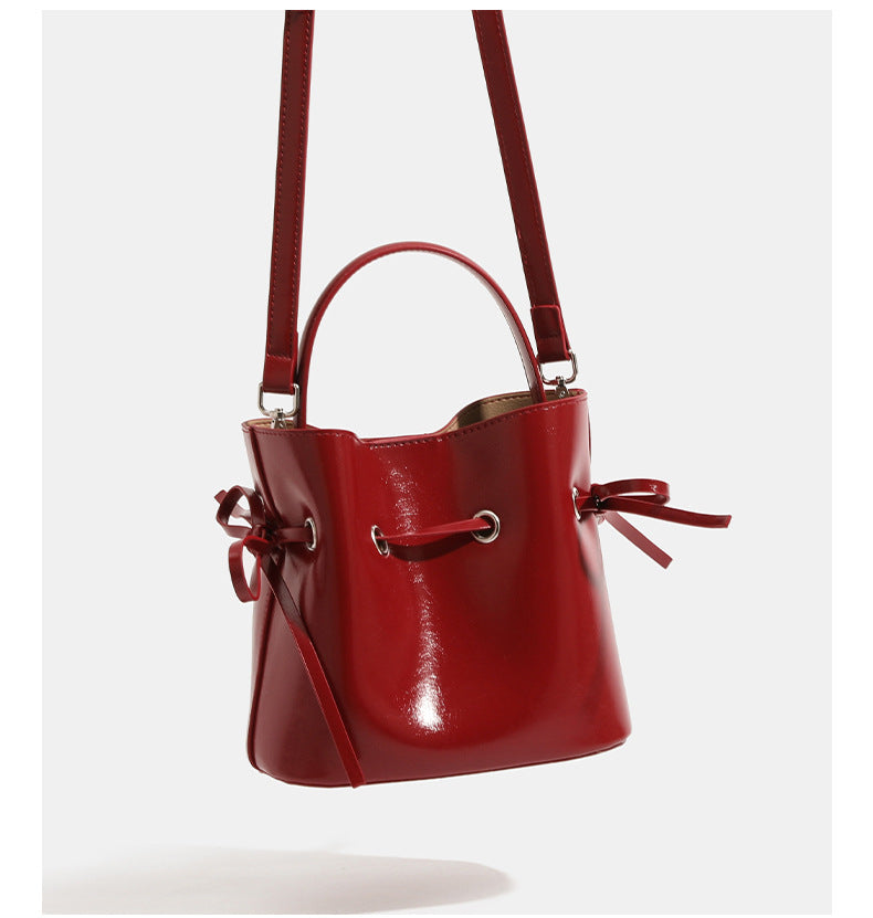Compact glossy leather-look bucket bag with drawstring bow-tie closure, rounded top handle, and silver-tone grommet hardware