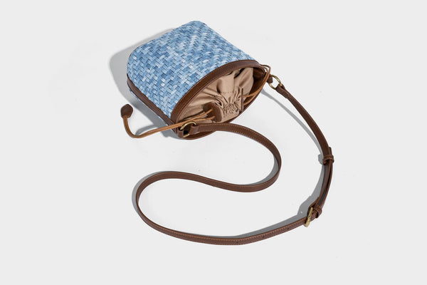 Small light blue woven bucket bag with brown leather-look trim, braided top handle, brass-tone ring hardware, drawstring