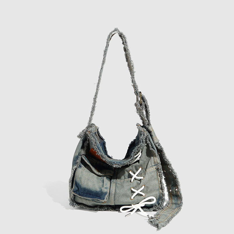Slouchy distressed denim shoulder bag with frayed edges, white lace-up cross detail, front snap pocket, and long shoulder