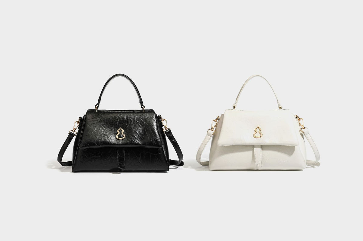 Structured black satchel bag with glossy crinkled leather-look finish, gold-tone emblem flap closure, slim top handle, and