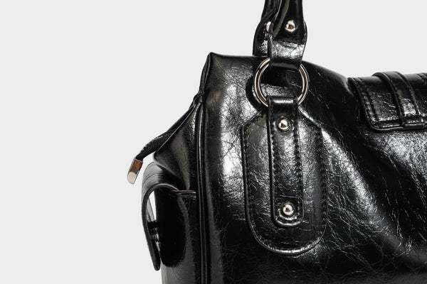 Glänzende, geknautschte Barrel-Satchel mit zwei oberen Tragegriffen, vorderem Klappen-Riemen-Panel, Ring- und Nietenbeschlägen sowie einem abnehmbaren