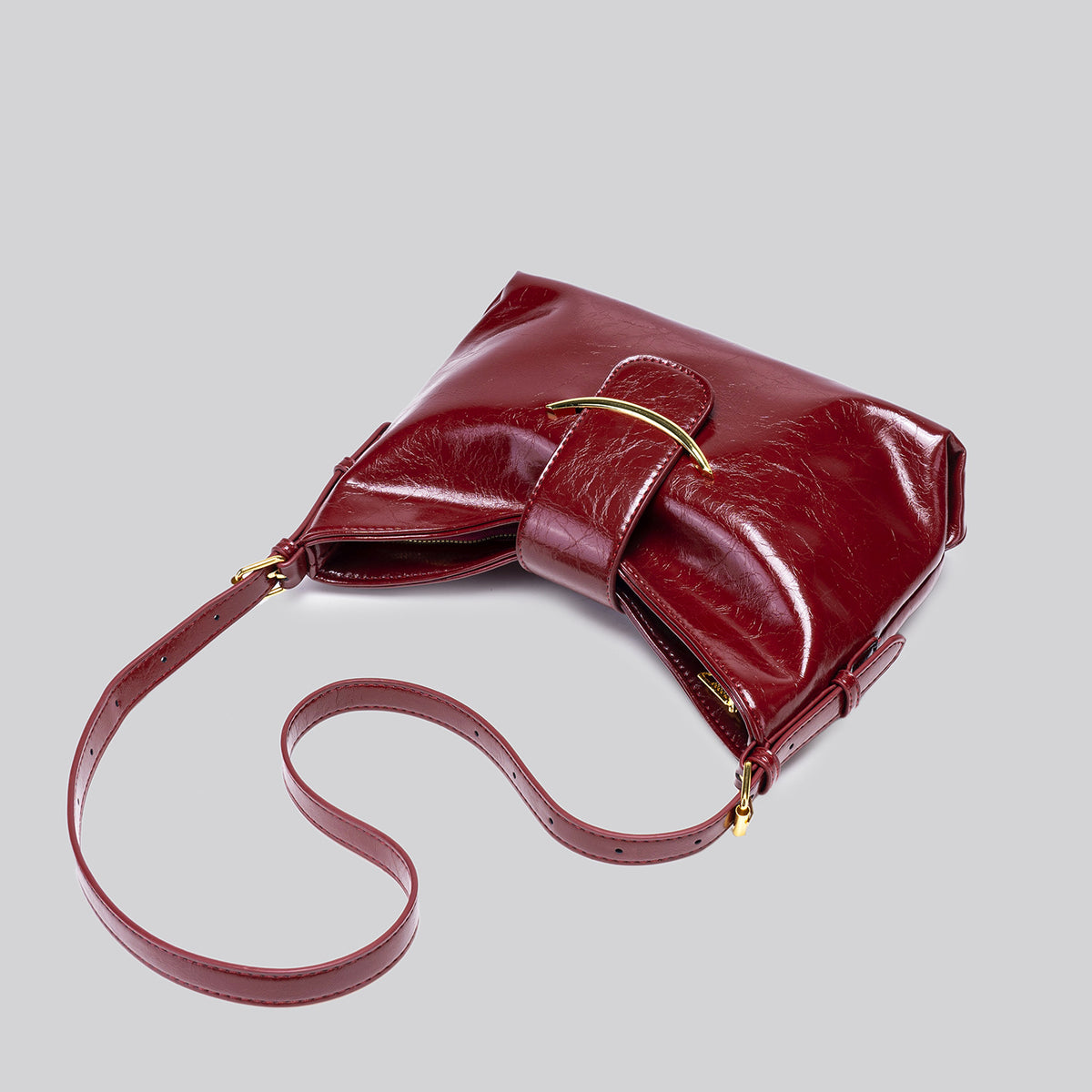 Glossy burgundy leather-look shoulder bag with curved gold-tone bar front closure, crinkled oil-wax texture, and adjustable