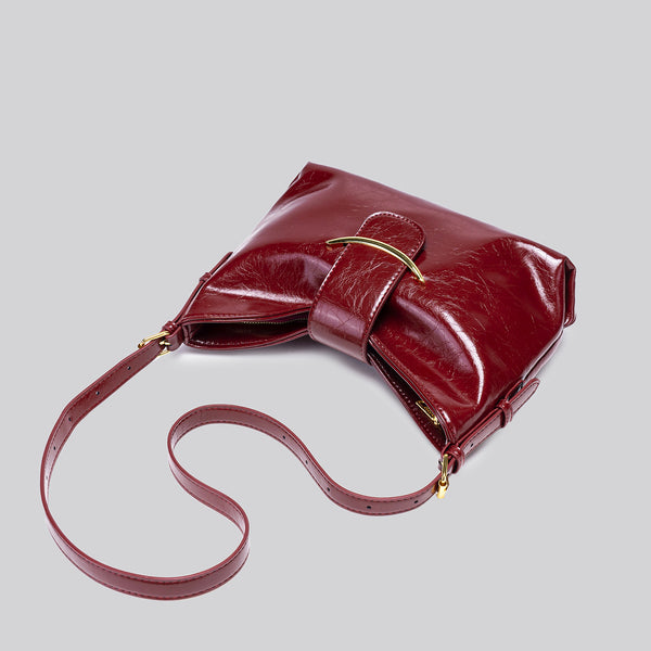 Glossy burgundy leather-look shoulder bag with curved gold-tone bar front closure, crinkled oil-wax texture, and adjustable