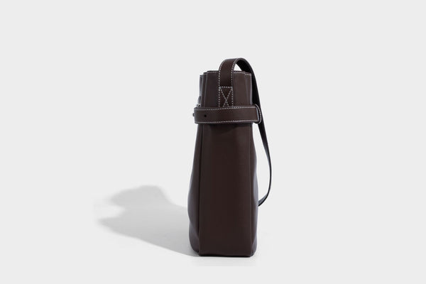 Structured dark brown leather-look bucket bag with white contrast stitching, front wraparound belt buckle strap, and long