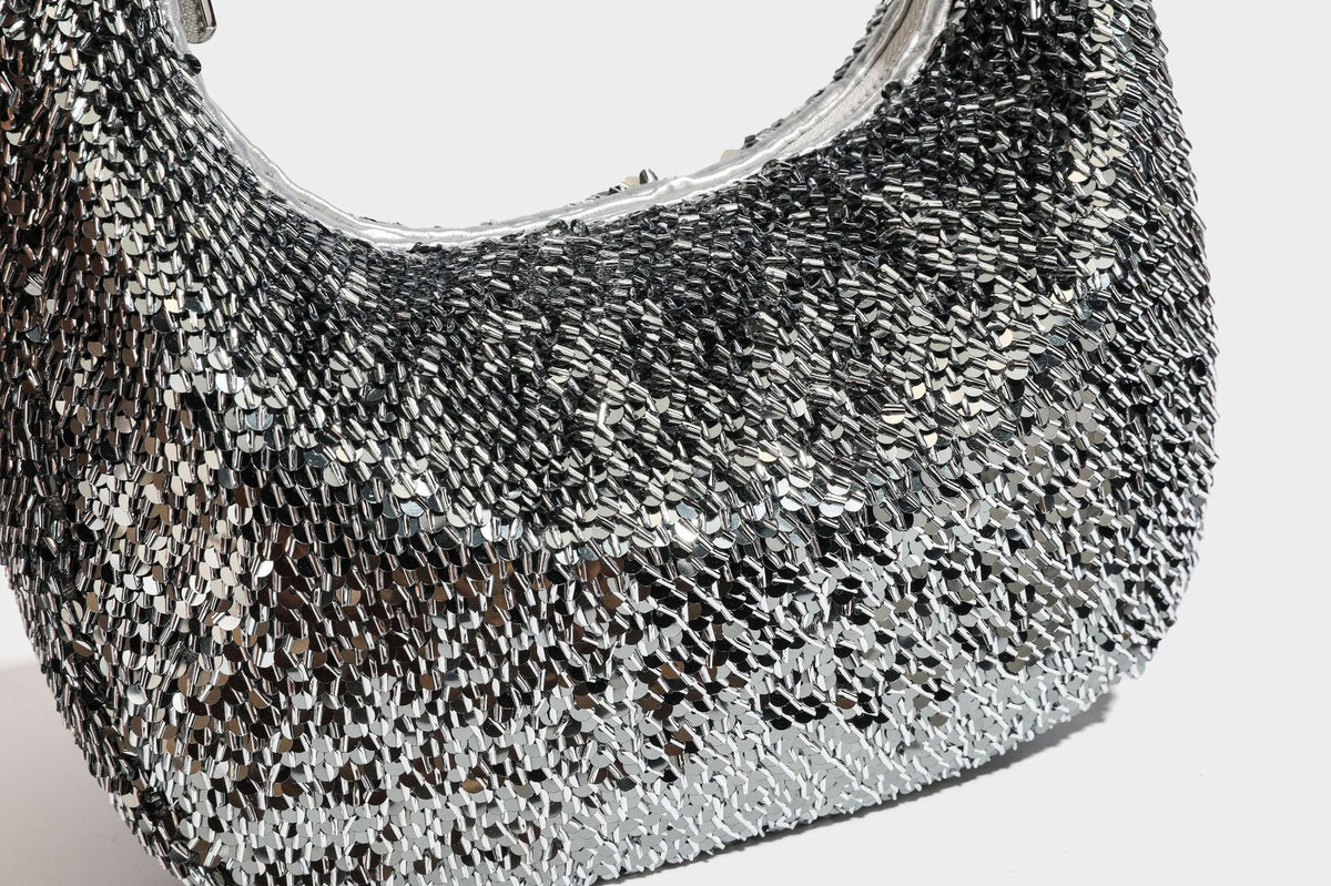 Kleine sichelförmige Schultertasche mit vollständig mit silberfarbenen Pailletten besetzter Außenseite, transparentem Henkel aus kugelförmigen Perlen und Reißverschluss in Silberoptik