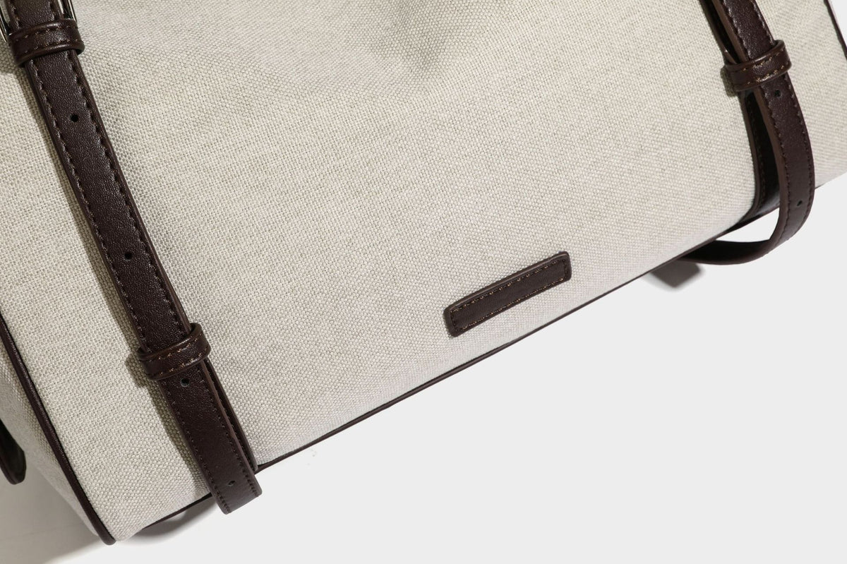 Beige canvas barrel bag with dark brown leather-look trim, silver-tone hardware, top zip closure, and adjustable shoulder