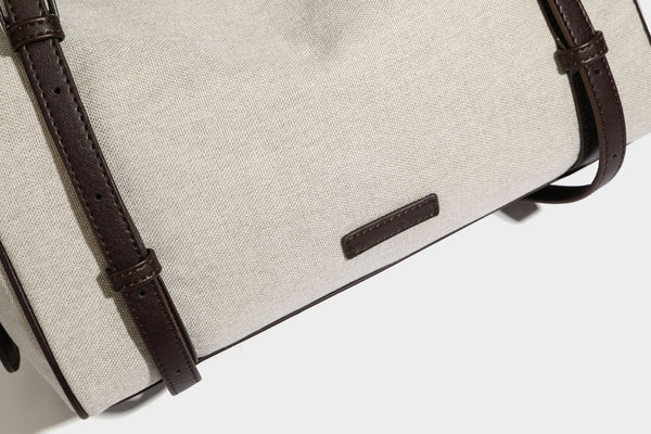 Beige canvas barrel bag with dark brown leather-look trim, silver-tone hardware, top zip closure, and adjustable shoulder