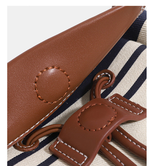 Cremefarbene Canvas-Beuteltasche mit horizontalen marineblauen Streifen, cognacfarbenem Besatz und Überschlag in Lederoptik, goldfarbenen Metalldetails und wandelbarem