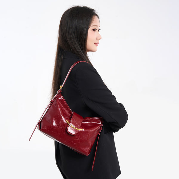 Glossy burgundy leather-look shoulder bag with curved gold-tone bar front closure, crinkled oil-wax texture, and adjustable