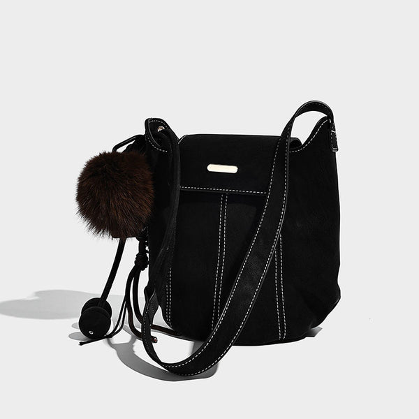 Small matte textured bucket crossbody bag with flap closure, gold-tone bar hardware, contrast stitching, and pompom tassel