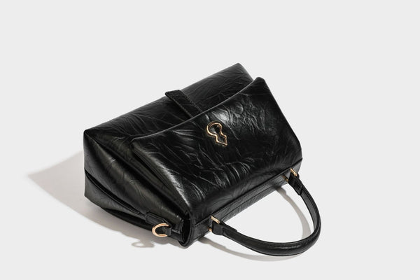 Structured black satchel bag with glossy crinkled leather-look finish, gold-tone emblem flap closure, slim top handle, and