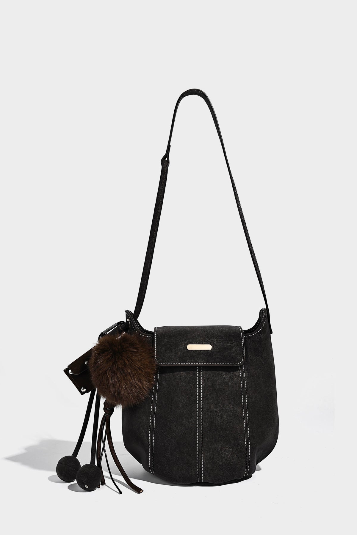 Small matte textured bucket crossbody bag with flap closure, gold-tone bar hardware, contrast stitching, and pompom tassel