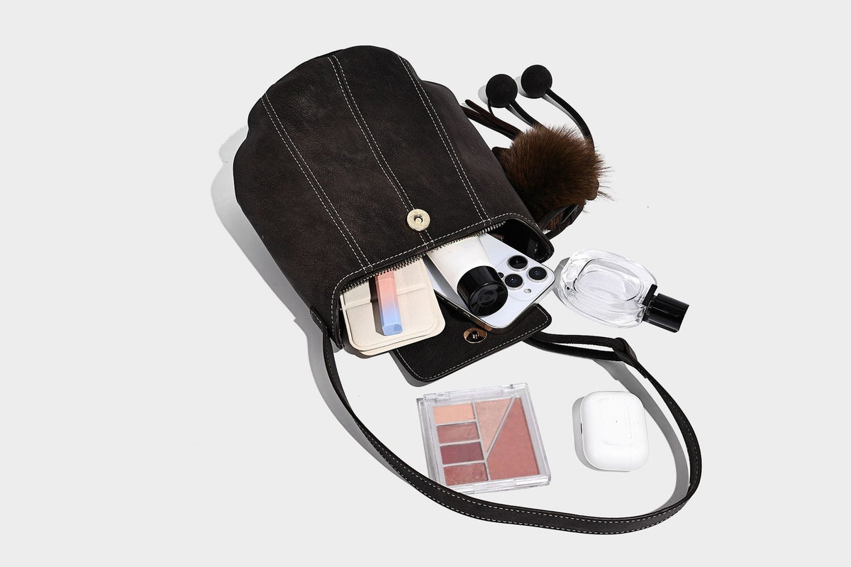 Small matte textured bucket crossbody bag with flap closure, gold-tone bar hardware, contrast stitching, and pompom tassel