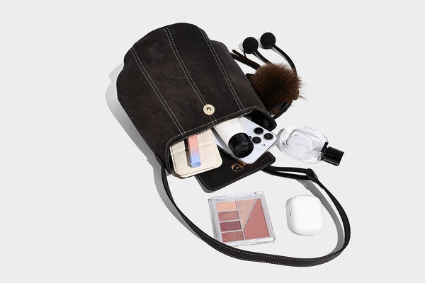 Small matte textured bucket crossbody bag with flap closure, gold-tone bar hardware, contrast stitching, and pompom tassel