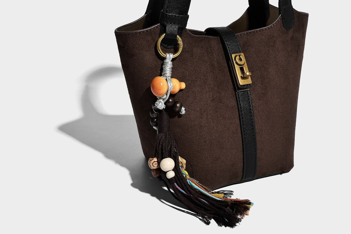 Small structured trapezoid tote bag in matte suede-look finish with black contrast trim, gold-tone front clasp, tassel charm