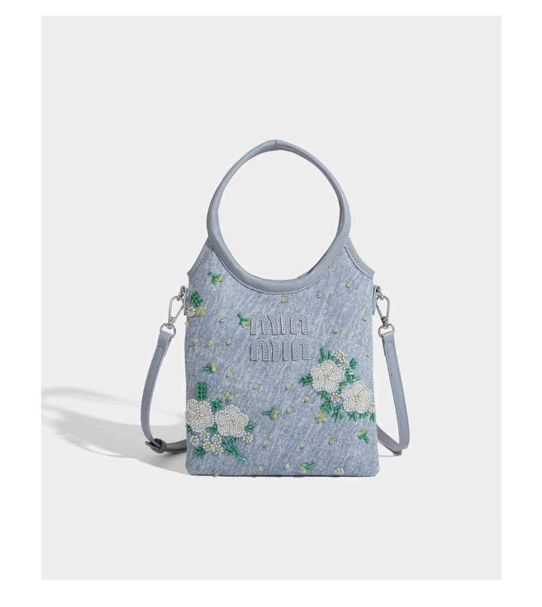 Kleine hellblaue Mini-Tote-Bag aus Stoff mit weißer Perlenblumenverzierung, runden Henkeln, silberfarbenen Beschlägen und