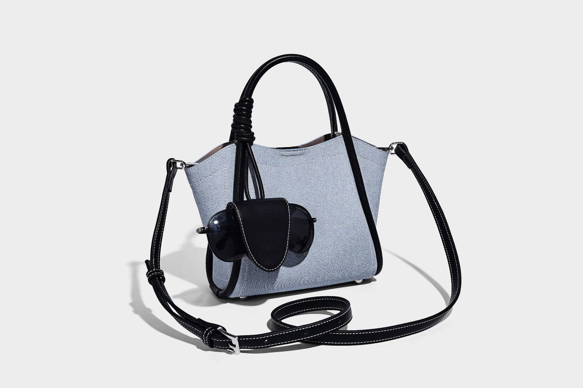 Light blue denim trapezoid tote bag with black leather-look trim, knotted top handles, detachable crossbody strap, front