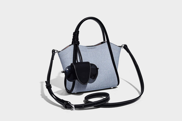 Light blue denim trapezoid tote bag with black leather-look trim, knotted top handles, detachable crossbody strap, front