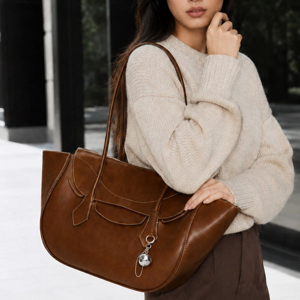 Large winged leather-look tote bag with single arched handle, stitched flap-front panel, and silver ball charm in cognac